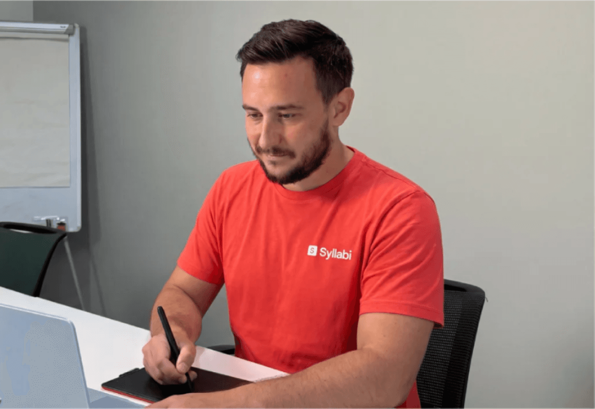A Syllabi teacher in a branded T-shirt working at a desk with a graphics tablet and pen
